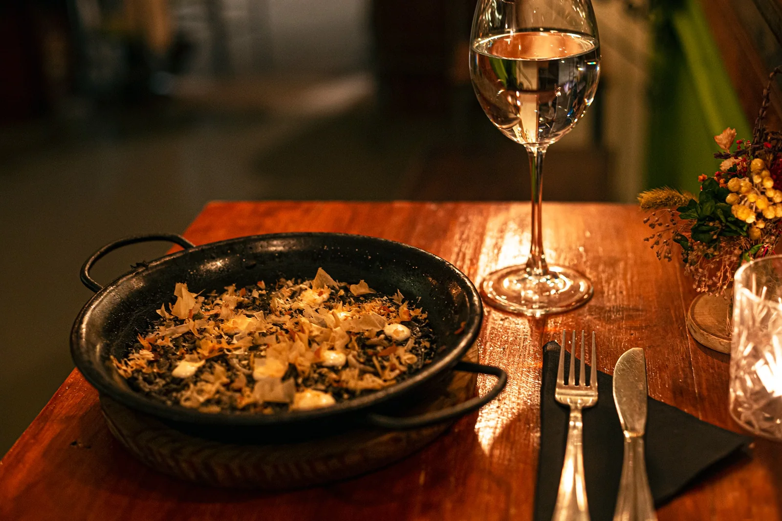 Una paella de arroz oscuro y una copa de vino blanco sobre una mesa de madera a la luz de las velas