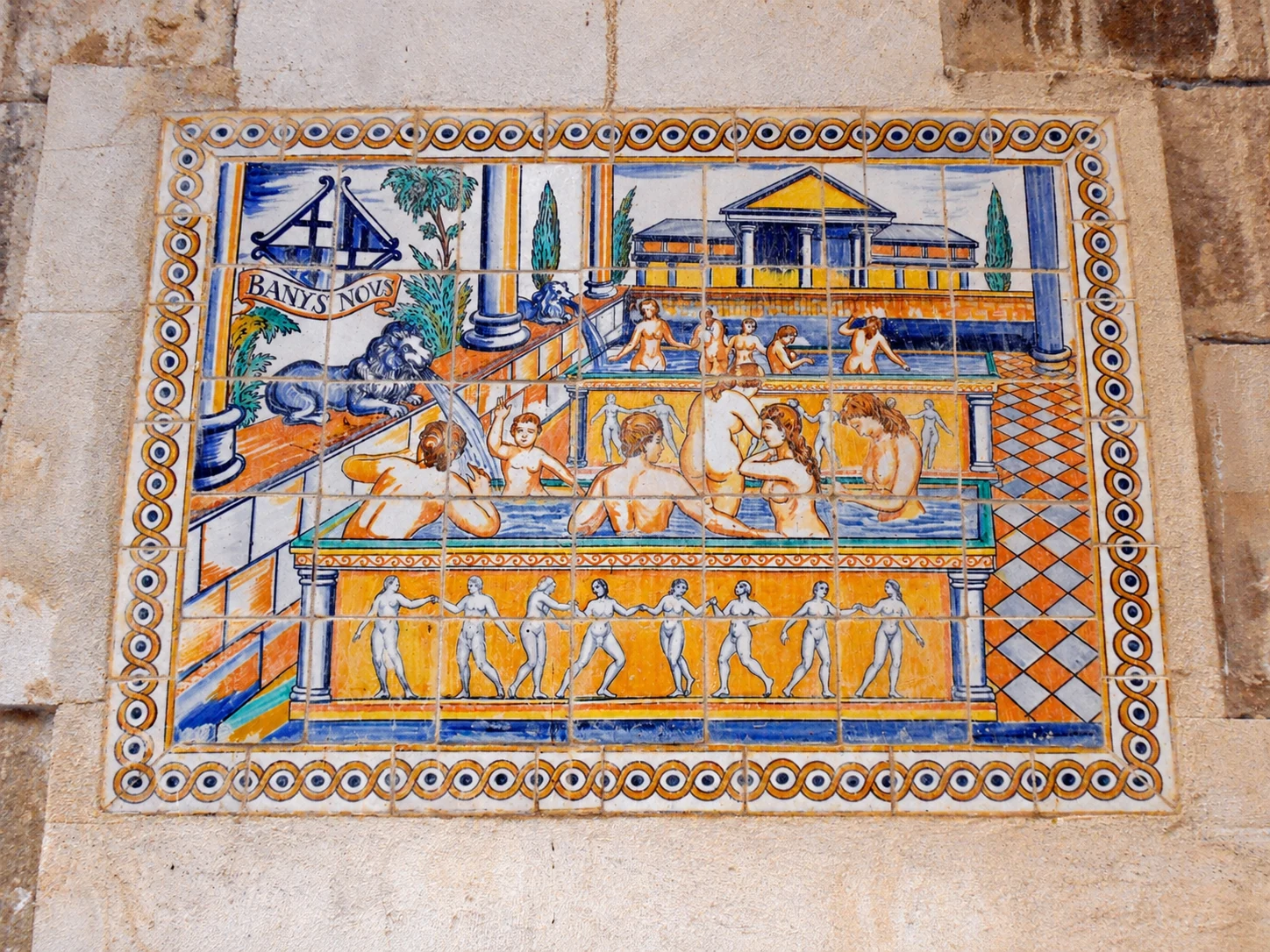 El mural de cerámica de Banys Nous sobre una pared de piedra: bañistas en estanques entre columnas, una fuente con cabeza de león y la inscripción «Banys Nous», en azul, amarillo y naranja
