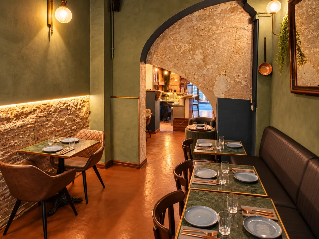 El comedor del fondo: paredes verde oliva, un muro de piedra iluminado, un largo banco de piel y mesas puestas, con un arco que da a la barra