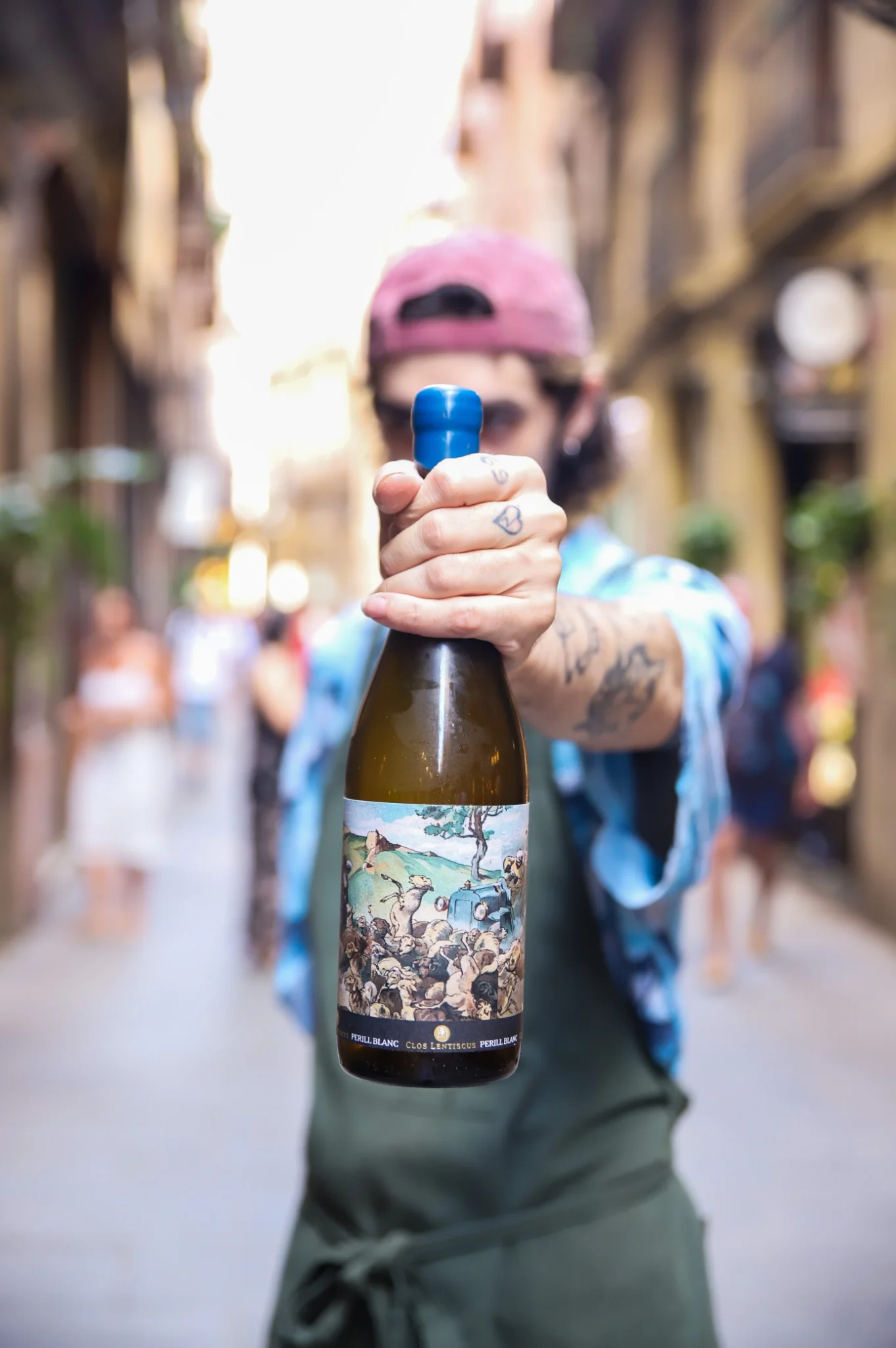 Una persona con delantal verde y gorra sostiene hacia la cámara una botella de vino blanco Perill Blanc de Clos Lentiscus, en una calle estrecha del casco antiguo